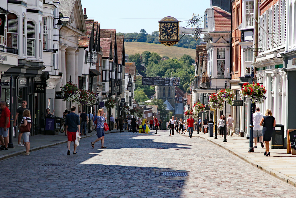 Guildford, Surrey UK - August 3, 2018. Bustling, busy high street lined with shops and stores with people buying things and with the local landmark of the guildhall town clock above the street